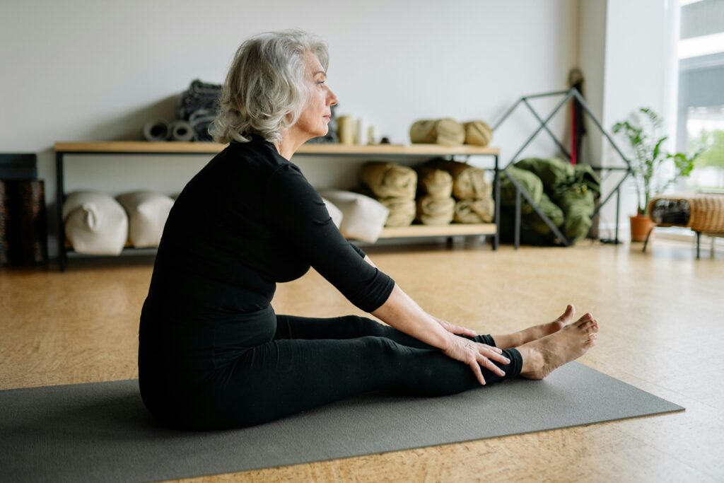 Senior woman in black clothing doing yoga stretching on a yoga mat indoors.