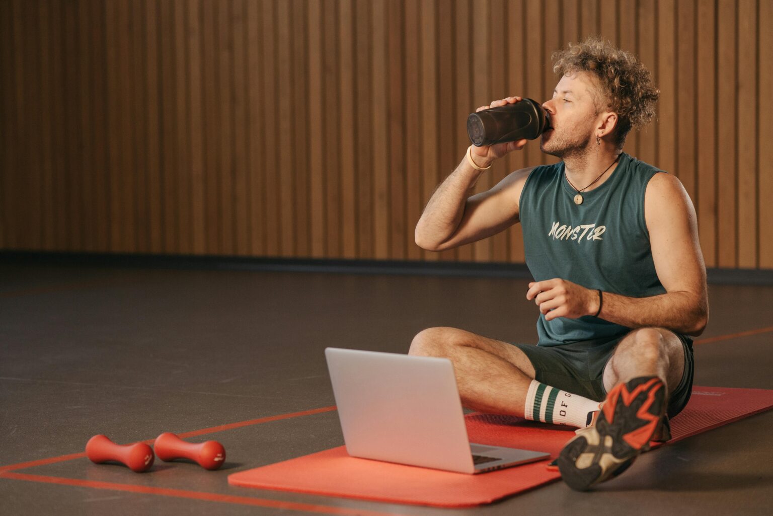 Man in gym drinking water during workout on exercise mat with laptop.